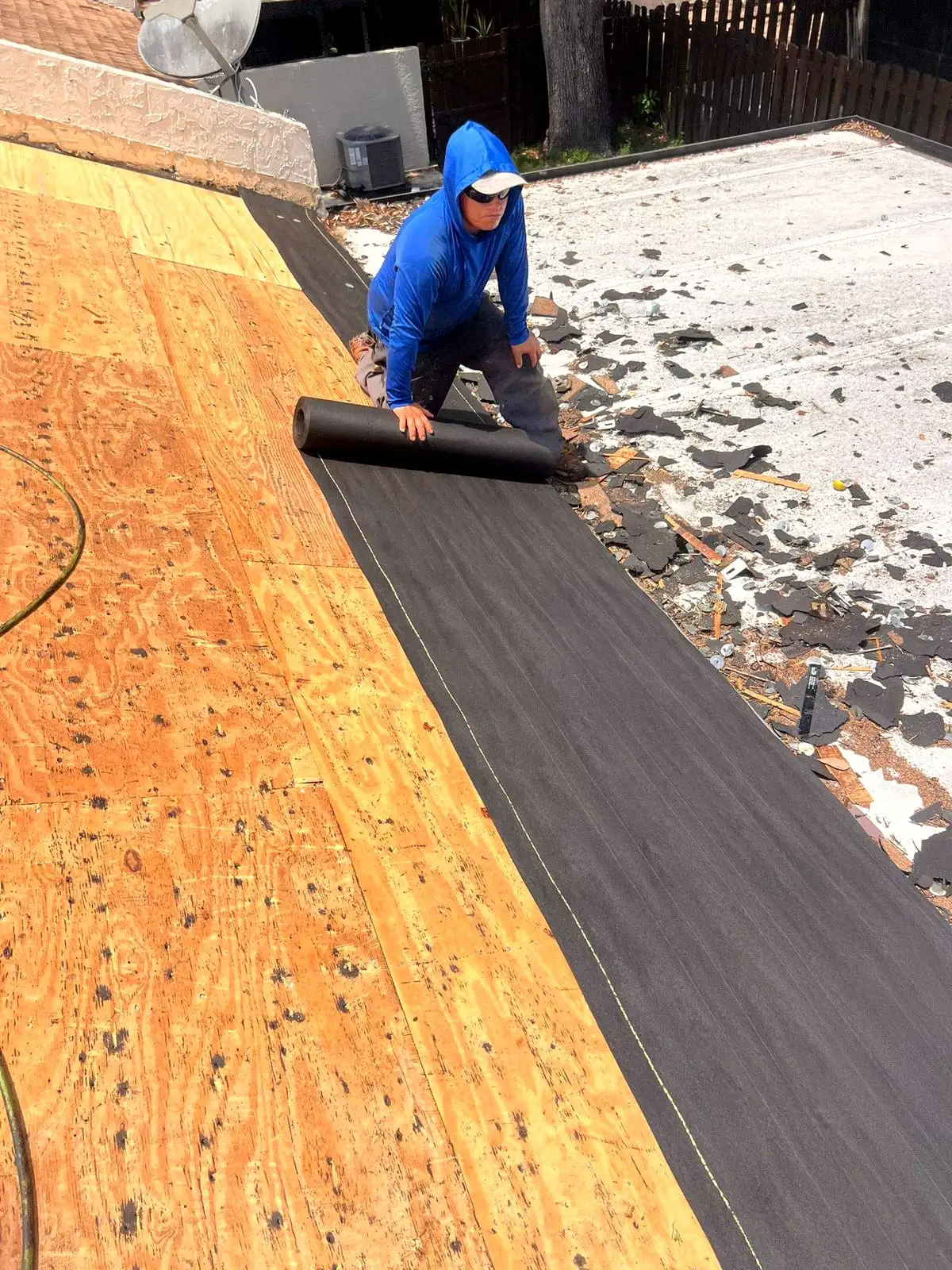 Roofer rolling underlayment over new plywood decking, with tear-off debris on the roof beside it